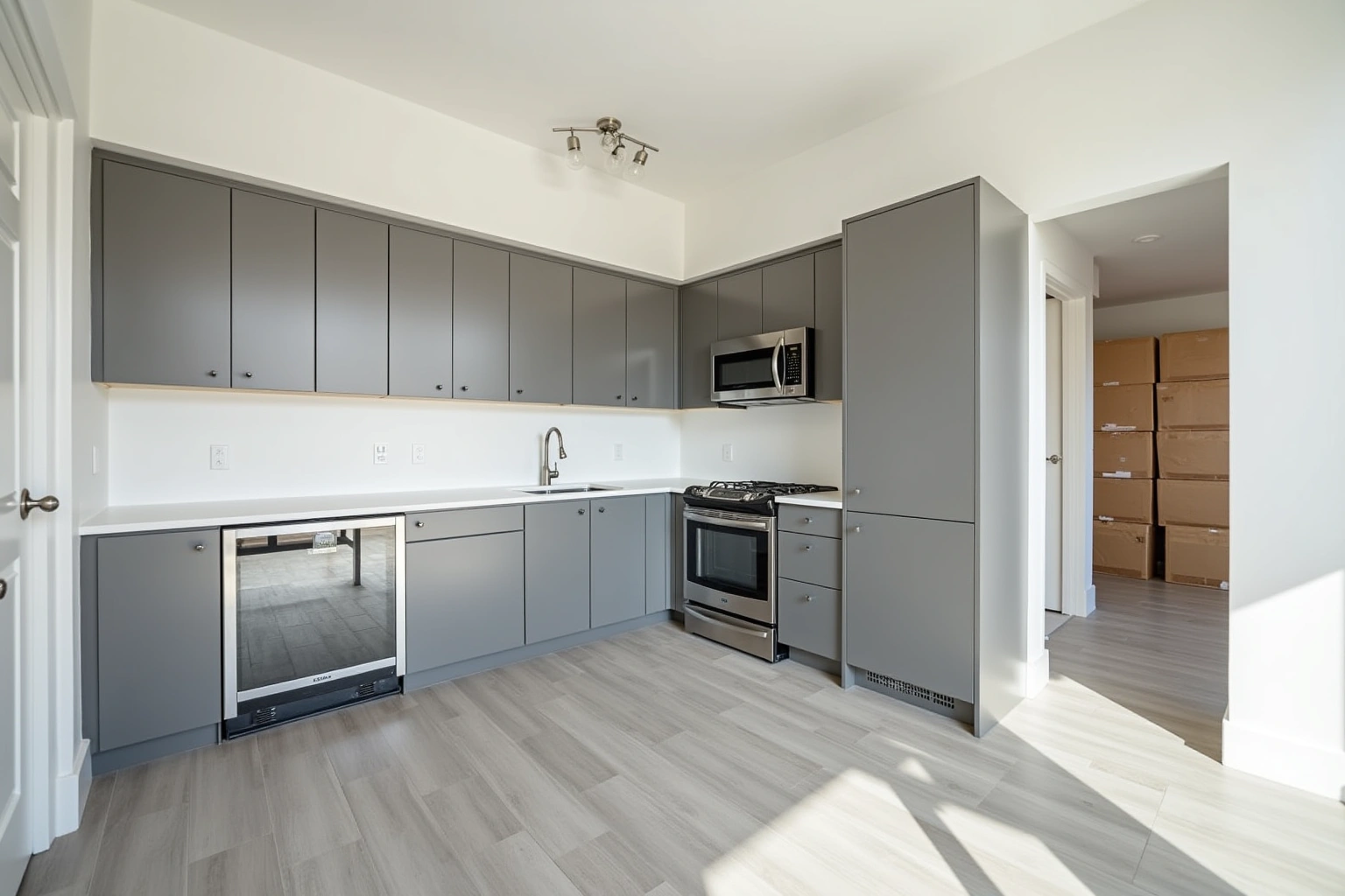 Flat-panel grey apartment kitchen with white quartz counters, stainless appliances and stacked cartons through the doorway.