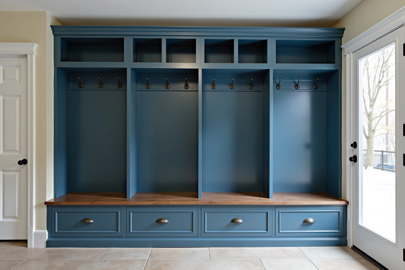 Four deep blue mudroom lockers with hooks, open cubbies above and a wood bench over drawers, beside a glass door.
